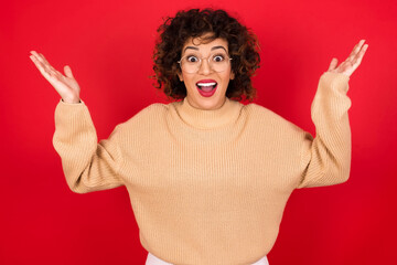 Young beautiful Arab woman wearing beige sweater against red background raising hands up, having eyes full of happiness rejoicing his great achievements. Achievement, success concept.