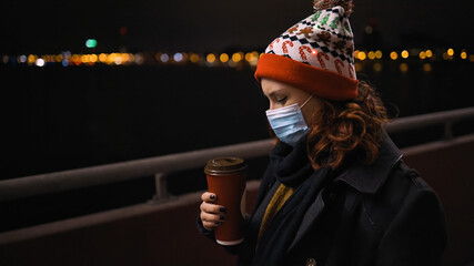 Side view: Tired Woman in medical mask and Xmas hat holds coffee cup