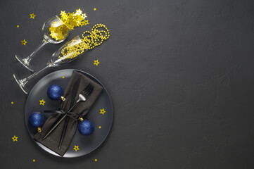 Festive table setting with black plate, cutlery, wine glasses with gold confetti and blue xmas balls on dark tabletop. Christmas table setting. Flat lay, top view. Minimal style, copy space