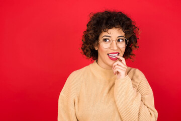 Young beautiful Arab woman wearing beige sweater against red background with thoughtful expression, looks to the camera, keeps hand near face, bitting a finger thinks about something pleasant.