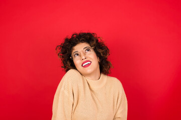 Portrait of mysterious Young beautiful Arab woman wearing beige sweater against red background looking up with enigmatic smile. Advertisement concept.
