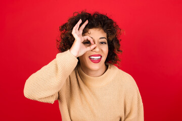 Young beautiful Arab woman wearing beige sweater against red background with happy face smiling doing ok sign with hand on eye looking through finger.