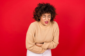 Fototapeta na wymiar Young beautiful Arab woman wearing beige sweater against red background suffering from strong stomachache.