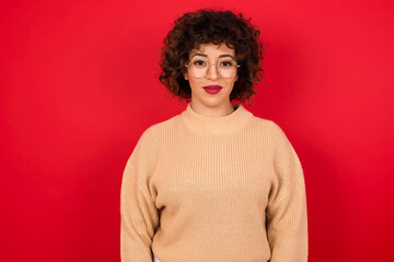 Fototapeta na wymiar Happy Young beautiful Arab woman wearing beige sweater against red background looking at camera with charming cute smile.