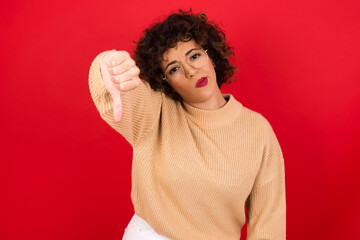Fototapeta na wymiar Discontent Young beautiful Arab woman wearing beige sweater against red background shows disapproval sign, keeps thumb down, expresses dislike, frowns face in discontent. Negative feelings.