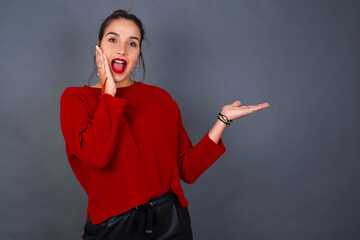 Crazy Young beautiful brunette woman wearing red sweater against gray background advising discount prices hold open palm new product