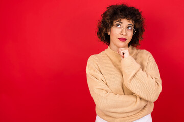 Fototapeta na wymiar Face expressions and emotions. Thoughtful young beautiful Arab woman wearing beige sweater against red background holding hand under his head, having doubtful look.