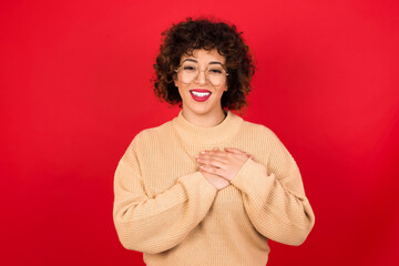 Honest Young beautiful Arab woman wearing beige sweater against red background keeps hands on...