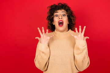 Joyful excited lucky Young beautiful Arab woman wearing beige sweater against red background cheering, celebrating success, screaming yes with clenched fists