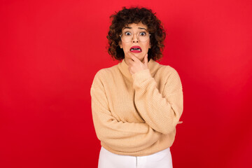 Fototapeta na wymiar Young beautiful Arab woman wearing beige sweater against red background covering mouth with hands scared from something or someone bitting nails