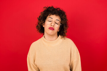 Fototapeta na wymiar Gloomy, bored Young beautiful Arab woman wearing beige sweater against red background frowns face looking up, being upset with so much talking hands down, feels tired and wants to leave.
