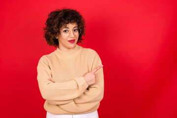 Fototapeta na wymiar Young beautiful Arab woman wearing beige sweater against red background smiling broadly at camera, pointing fingers away, showing something interesting and exciting.