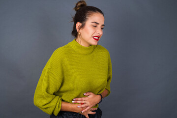 Young beautiful hispanic woman wearing green sweater against gray background with hand on stomach...