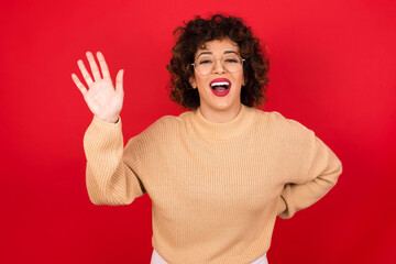 Young beautiful Arab woman wearing beige sweater against red background waiving saying hello or goodbye happy and smiling, friendly welcome gesture.