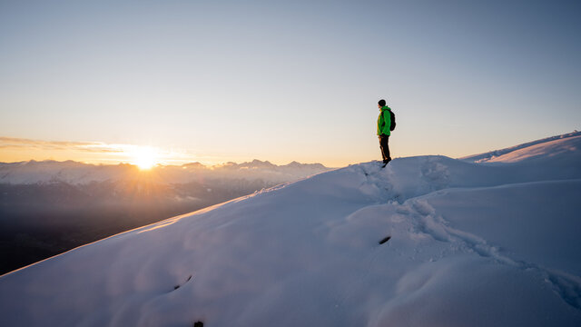 Winter Trekker Staying On Mountain Peak He Climbed And Enjojing Wide Panorame View Of Valley During Sunrise. Active Winter Trekking Concept Image. Adventure Concept