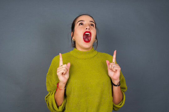 Young beautiful hispanic woman wearing green sweater against gray background amazed and surprised looking up and pointing with fingers and raised arms.