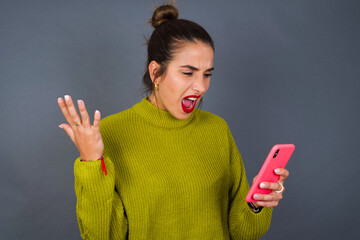 Angry Young beautiful hispanic woman wearing green sweater against gray background screaming on the...