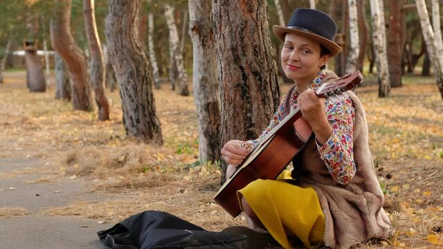 Beautiful woman in a hat playing acoustic guitar in park. Street musician performer sitting on ground performs a song asking for money.