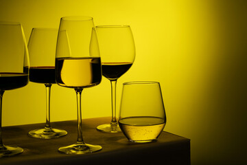 Different glass goblets with white and red wine stand against the yellow light.Glasses of different shapes and heights.Air bubbles are visible in the glasses. Sommelier.Space for text. Assorted wines.