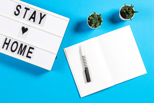 White lightbox with text STAY HOME and notebook on blue background. Online learning, working, social distancing concept