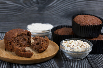 Oatmeal chocolate muffins. Drawn up on a silver platter. One is broken to show a cut. Nearby are cereals and oat flour. On black pine boards.