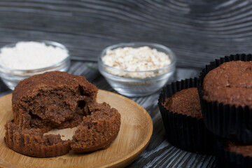 Oatmeal chocolate muffins. Drawn up on a silver platter. One is broken to show a cut. Nearby are cereals and oat flour. On black pine boards.