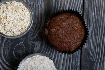 Oatmeal chocolate muffins. Drawn up on a silver platter. One is broken to show a cut. Nearby are cereals and oat flour. On black pine boards.