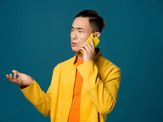 An Asian man in a yellow jacket is gesturing with his hands and talking on the phone