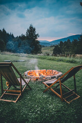 Campsite With Fire Pit And Two Empty Chairs. Burning Campfire with mountain landscape with evening sunset sky.