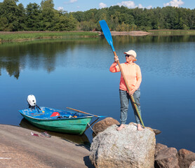 A happy woman with an oar stands on a rock on the shore of the lake next to the boat