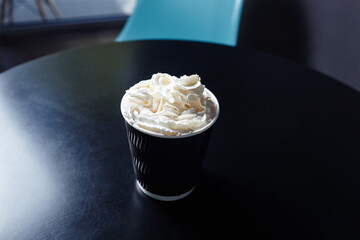 Disposable paper cup with tasty coffee drink with milk and whipped cream on wooden table in cafe. Selective focus, blurred background