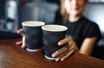 Fototapeta na wymiar Barista holds two disposable paper cups of coffee in coffee shop. Blurred image, selective focus