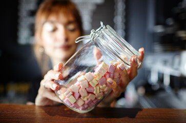 Barista holds jar with tasty mini marshmallows at bar counter in coffee shop. Blurred image,...