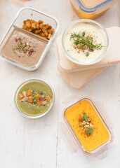 Batch cooking scene with four glass jars full of delicious different cream soups on a wooden table. Additional plastic food containers are piled.