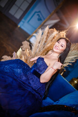 Beautiful woman in a blue ball gown sitting on sofa in blue room. dry flowers in a vase on the...
