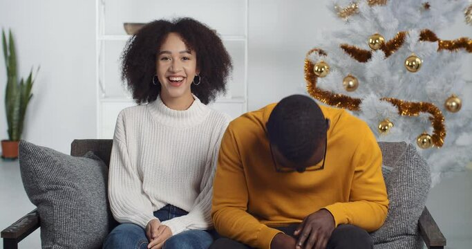 Couple afro american man husband boyfriend and black woman wife girlfriend sitting at home on couch in sweaters near Christmas tree laughing smiling with joke chatting online via video conference