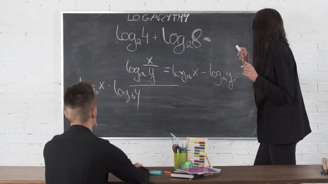 Education in high school and college. Young teacher in math lesson writes on a blackboard with chalk. Long loose black hair.