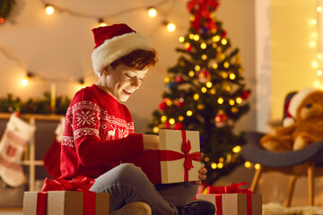 Fototapeta na wymiar Positive boy in red festive clothes sitting and unpacking boxes with Christmas presents