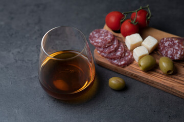 glass of whiskey and snacks sausage cheese olives and tomatoes on dark gray cement background