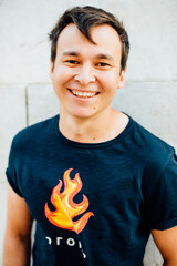 Close up portrait of young Asian man wearing black t-shirt with a symbol of fire and word "fire" written in Russian and smiling and looking at camera while standing against a white stone wall