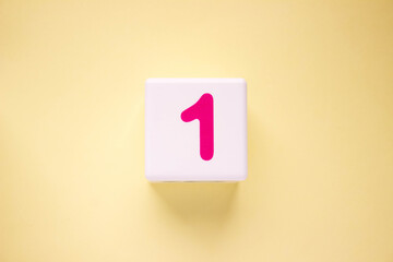Close-up photo of a white plastic cube with a pink number one on a yellow background. Object in the center of the photo