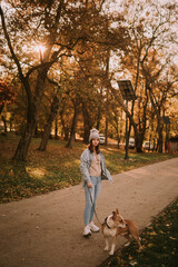 Fototapeta premium Full length of a beautiful teenage girl with a hat and casual dressed posing while holding the leash of her dog breed American Staffordshire Terrier. Autumn walk in the park