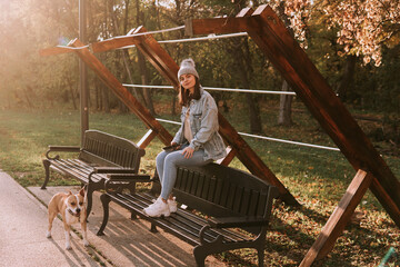 A cute young teenage girl with a hat in a denim coat is sitting on a park bench, and next to her is her brown dog breed American Staffordshire Terrier. Sunny autumn in the park