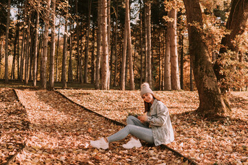 Fototapeta premium A smiling cute brunette caucasian girl with a hat, dressed in jeans sits in a park on the ground and uses the phone. Autumn walk in nature