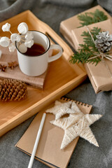 Obraz na płótnie Canvas Christmas background with mug of hot cocoa with marshmallow on wooden tray, Notepad with pencil, wrapped boxes and decoration. Cozy mood holiday. Hygge. Letter To Santa Claus concept. Selective focus