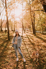 Fototapeta premium A young urban teenage girl with a hat in a denim coat standing in the park next to a brown American American Staffordshire Terrier. Autumn walk in nature