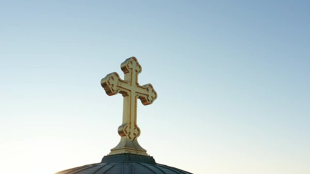 Silhouette of a golden cross against clear blue sky. Orthodox christian crucifix back illuminated by sunlight. Religious symbol of the faith in god made of gold. Aerial view. 