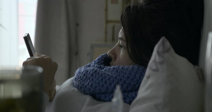 Sick Woman Using Smartphone Browsing Internet Tapping Touch screen and Coughing while Lying on Bed in front of Window in Bedroom 4K