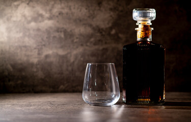 a bottle of whiskey in a square shape in the rays of light next to an empty glass on a dark gray cement background