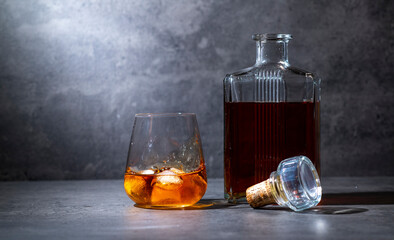 square whiskey bottle and glass with ice on dark gray cement background